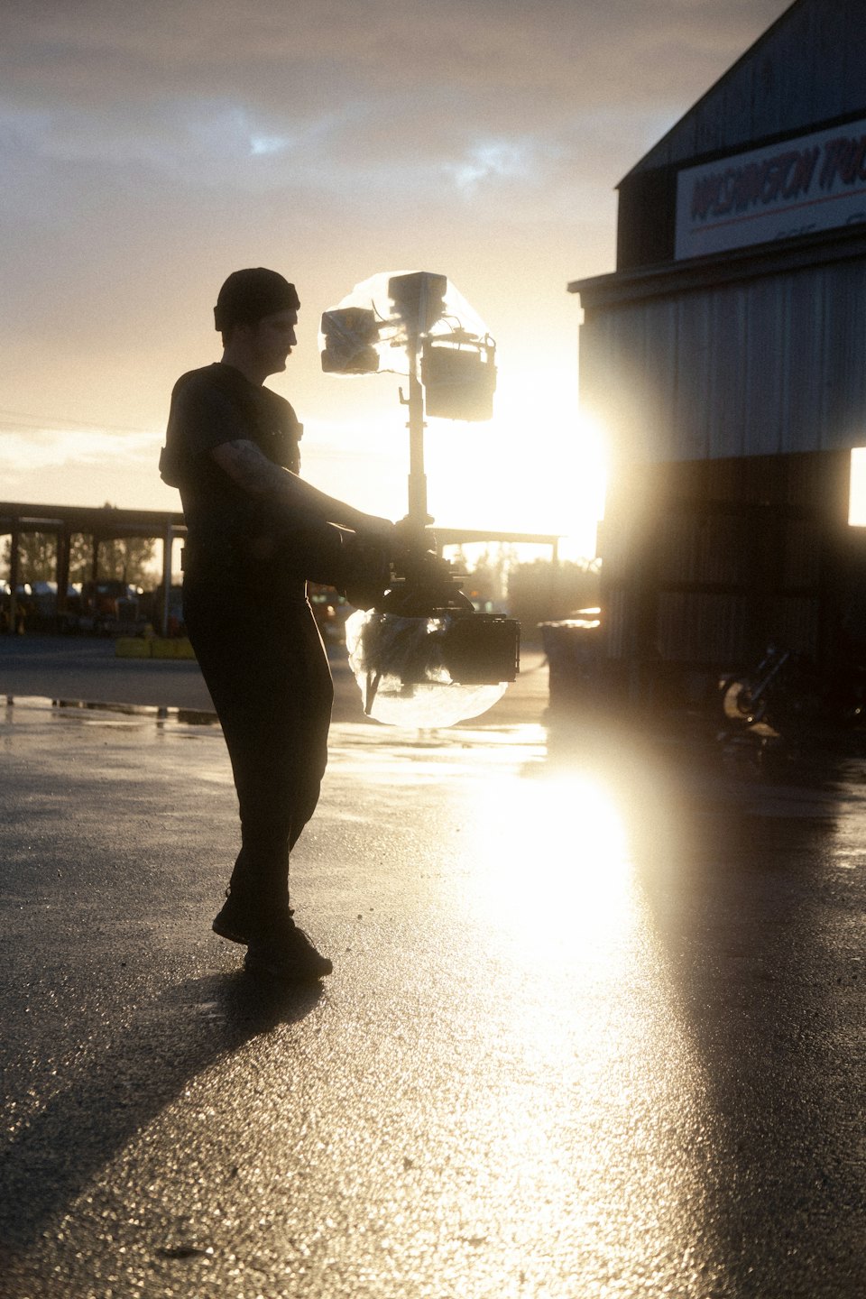 Film crew member silhouetted operating camera gimbal on wet asphalt.